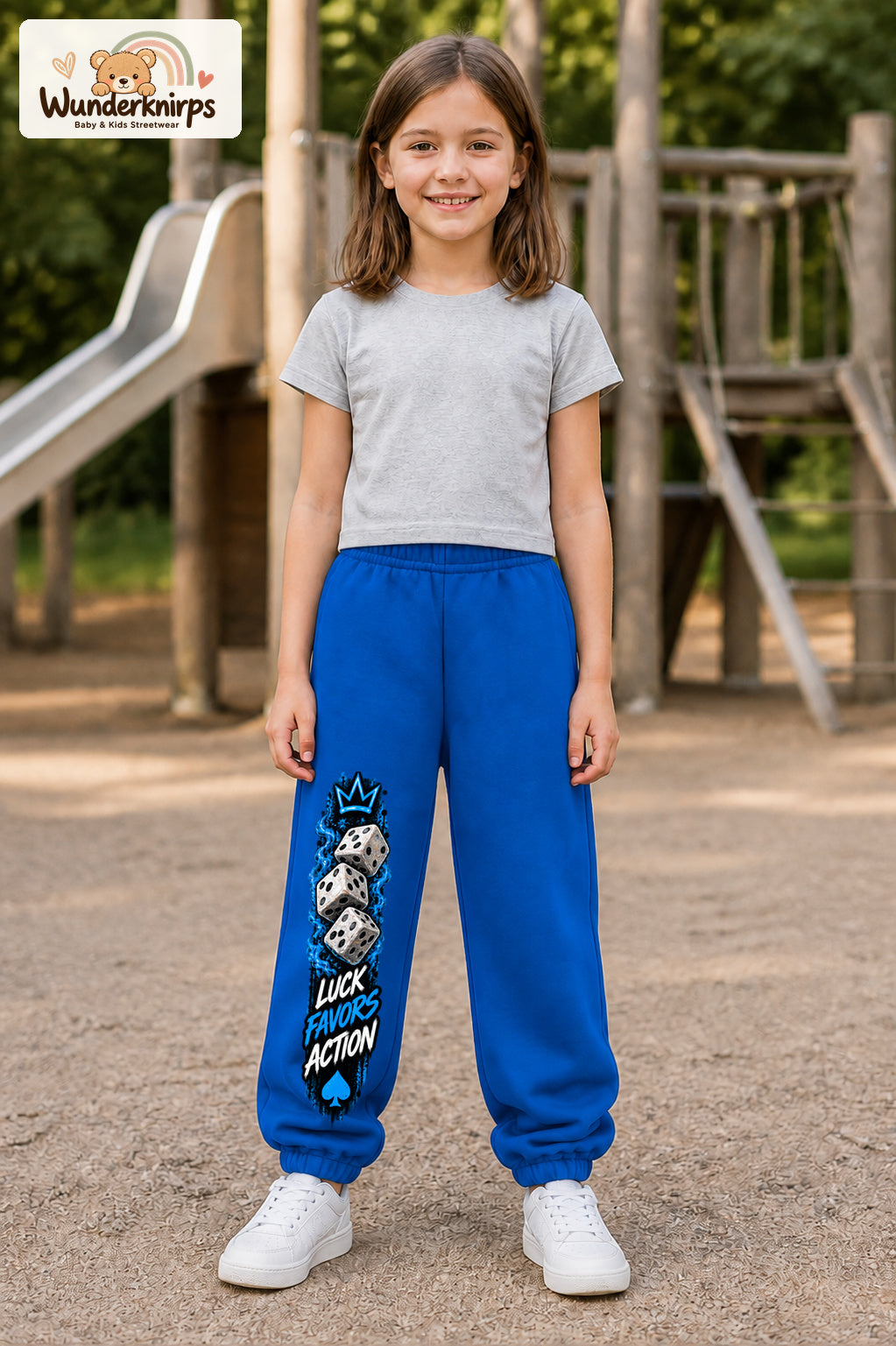 Mädchen trägt blaue Jogginghose mit Luck Favors Würfel-Print, von vorn auf dem Spielplatz.