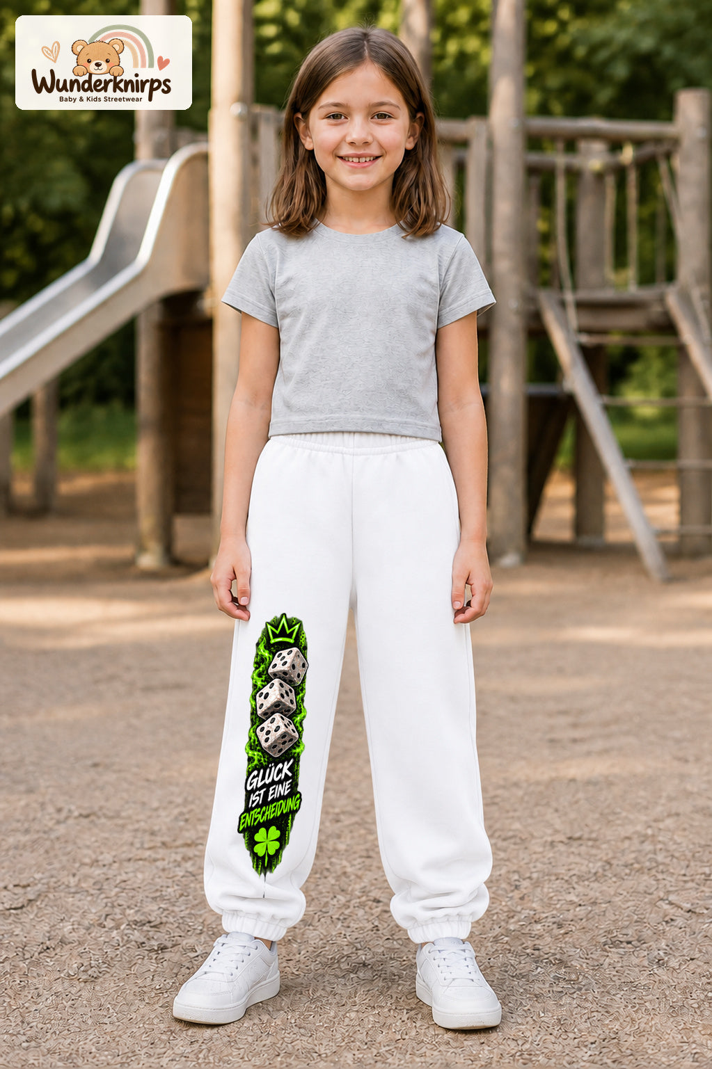 Mädchen trägt weiße Jogginghose mit Glück ist Entscheidung-Print, von vorn auf dem Spielplatz.