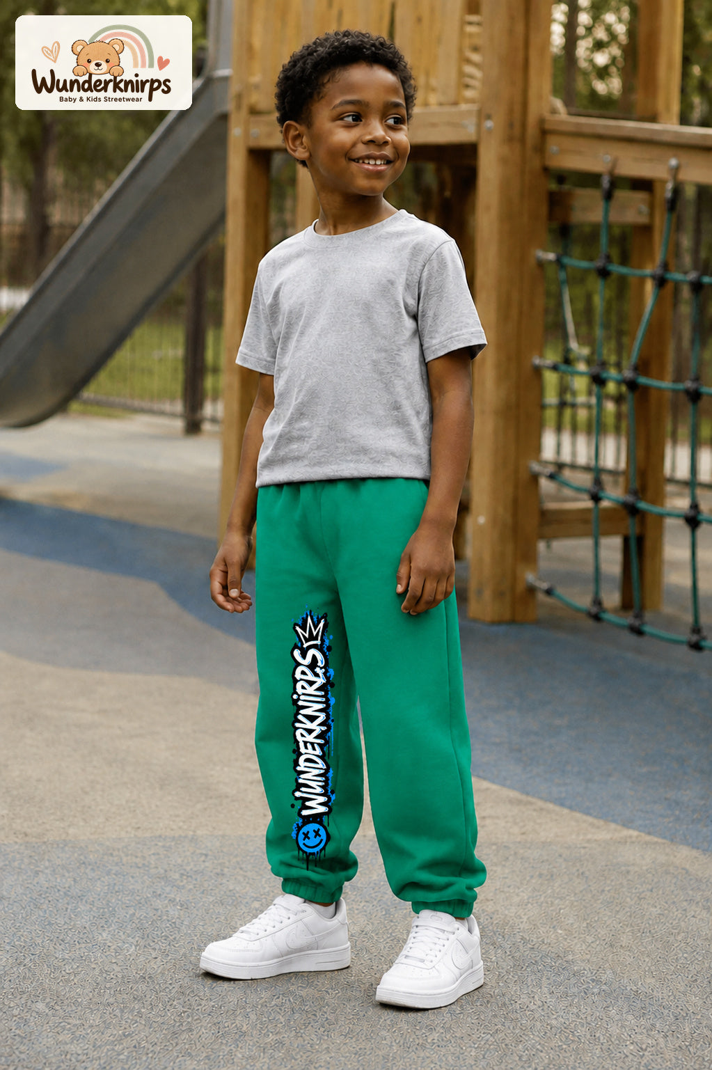 Junge trägt grüne Jogginghose mit Wunderknirps Graffiti-Print, leicht seitlich auf dem Spielplatz.