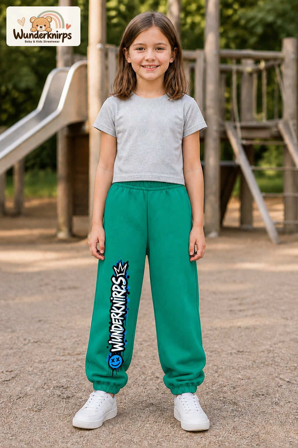 Mädchen trägt grüne Jogginghose mit Wunderknirps Graffiti-Print, von vorn auf dem Spielplatz.