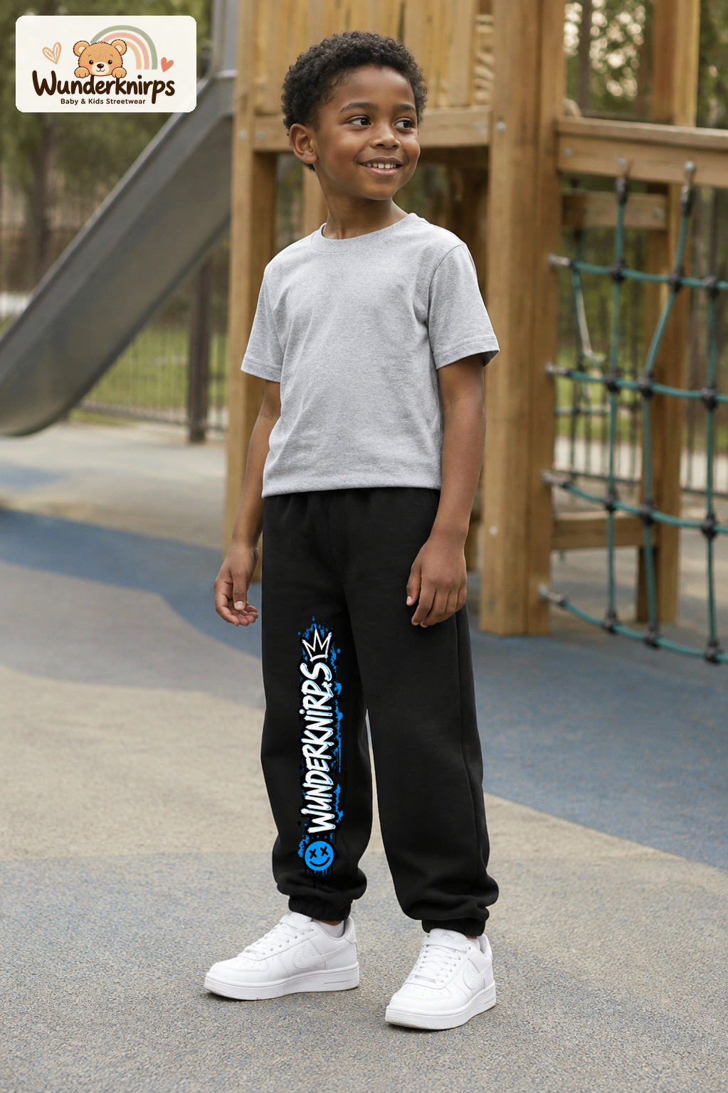 Junge trägt schwarze Jogginghose mit Wunderknirps Graffiti-Print, leicht seitlich auf dem Spielplatz.